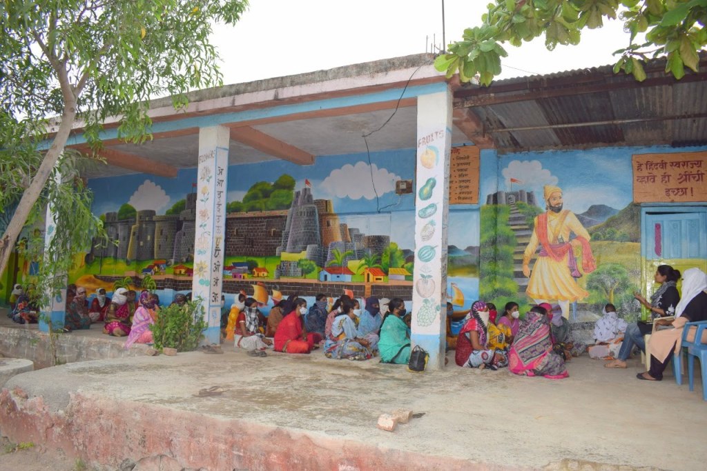 A large group of village women wearing masks sitting on an outdoor porch with vibrant murals, participating in a 
community wellness session led by Savine Ventures